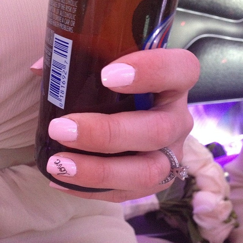 May include: A hand with pink nail polish and a ring on the ring finger holds a brown glass bottle. The bottle has a barcode and the text "love" is written on the finger.