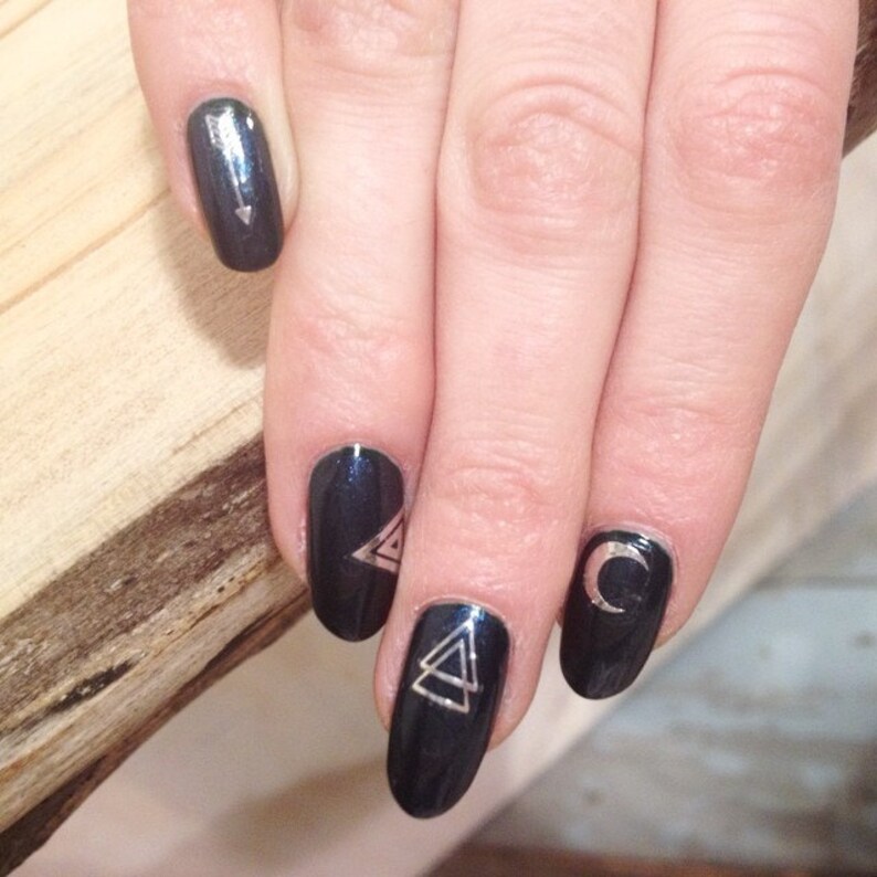 May include: Close-up of a hand with dark blue nail polish and silver geometric designs. The designs include a crescent moon, a triangle, and an arrow.