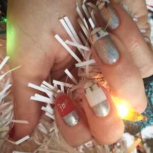 May include: Close-up of a hand with several fingernails painted with a white base and a silver glitter top coat. One fingernail has a red base with the word "love" written in white. Another fingernail has a white base with a design of a string of Christmas lights.