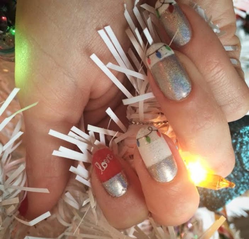 May include: Close-up of a hand with several fingernails painted with a white base and a silver glitter top coat. One fingernail has a red base with the word "love" written in white. Another fingernail has a white base with a design of a string of Christmas lights.