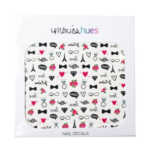 May include: A sheet of nail decals with a white background and a repeating pattern of black and pink designs. The designs include hearts, Eiffel Towers, rings, bowties, infinity symbols, and the words "love" and "je t'aime". The sheet is packaged in a white cardboard box with the brand name "monicahues" and the words "NAIL DECALS" printed on it.