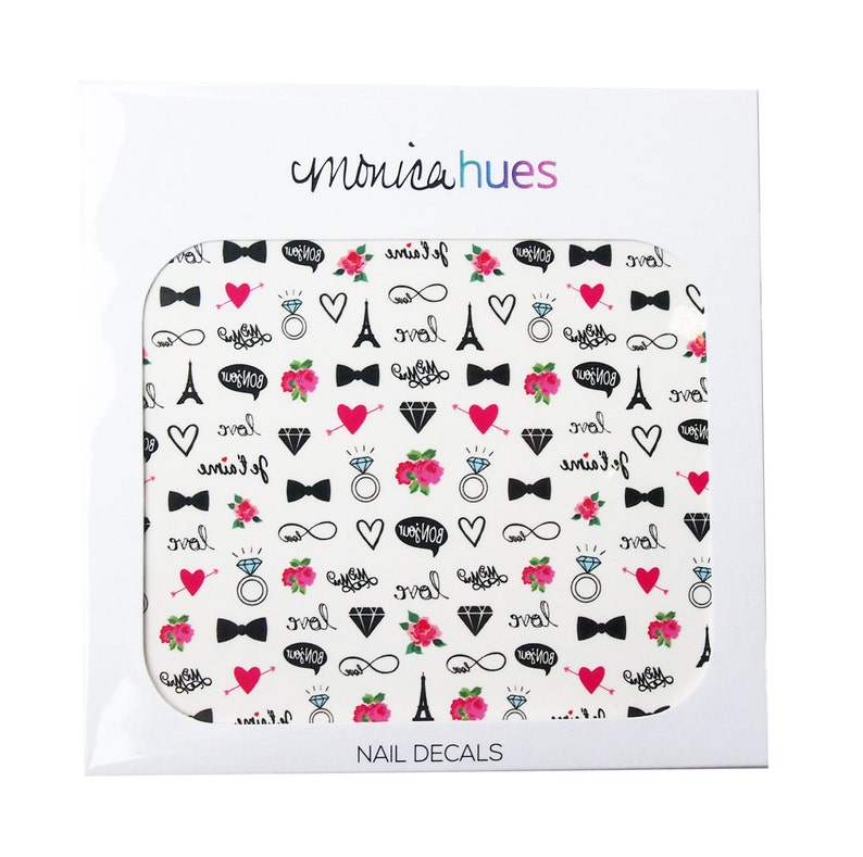 May include: A sheet of nail decals with a white background and a repeating pattern of black and pink designs. The designs include hearts, Eiffel Towers, rings, bowties, infinity symbols, and the words "love" and "je t'aime". The sheet is packaged in a white cardboard box with the brand name "monicahues" and the words "NAIL DECALS" printed on it.