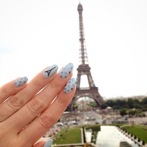 May include: A hand with light blue nail polish with white polka dots and one nail with a black Eiffel Tower design. The Eiffel Tower is in the background.