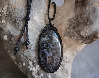 May include: A black macrame necklace featuring an oval pendant with a dark, mottled pattern of blues and grays. The pendant is framed by a detailed macrame border. The necklace has an adjustable cord with black beads.