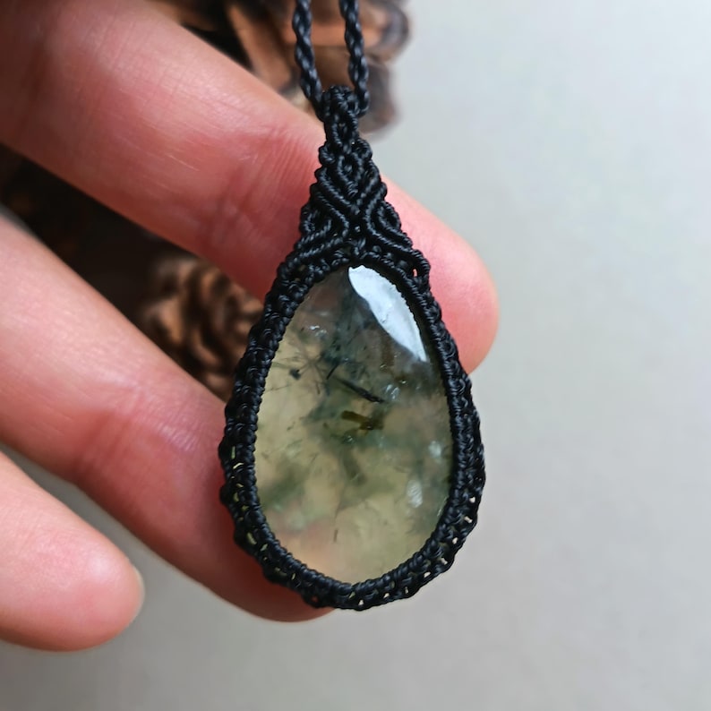 Op de afbeelding: Een druppelvormige hangerketting met een doorschijnende, lichtgroene steen met donkere insluitsels. De steen is gevat in een zwarte macrame-omgeving en hangt aan een bijpassend zwart koord. Een uniek sieraad.