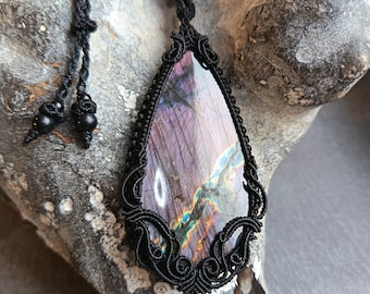 May include: A handcrafted macrame necklace featuring a large, teardrop-shaped labradorite stone. The stone displays a vibrant play of color, with hues of purple, blue, and green. The black macrame setting adds a contrasting frame to the iridescent gemstone.