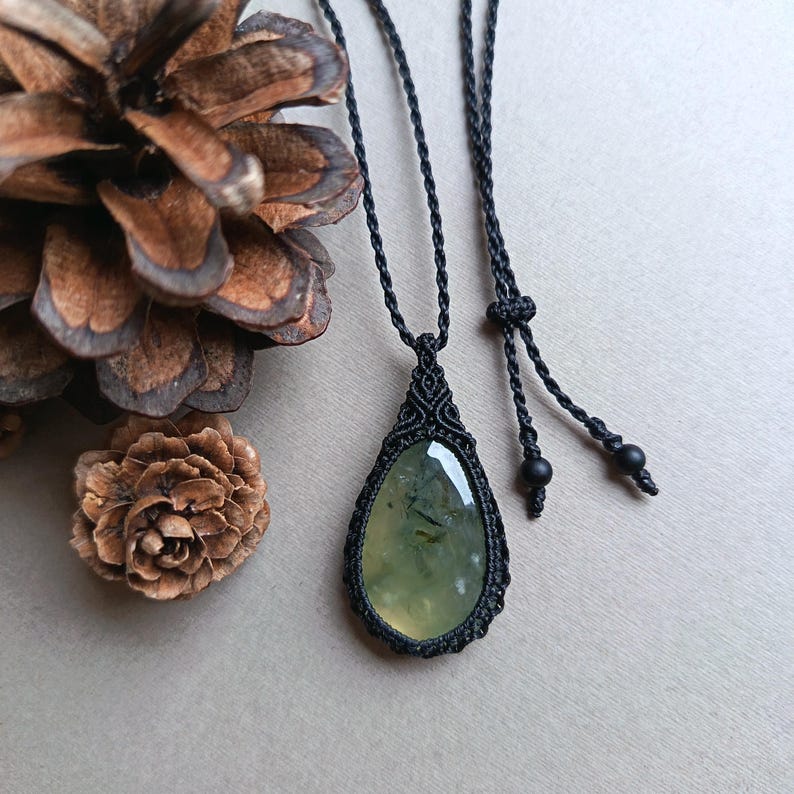 Op de afbeelding: Een handgemaakte macram&eacute; ketting met een druppelvormige, lichtgroene edelsteen hanger. De hanger is gevat in ingewikkeld zwart macram&eacute;, hangend aan een zwart koord. De ketting wordt getoond tegen een neutrale achtergrond met dennenappels.