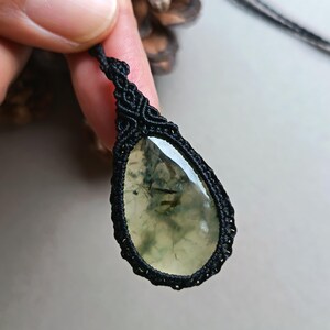 Op de afbeelding: Een druppelvormige hanger met een doorschijnende, lichtgroene steen, met donkergroene insluitsels. De steen is gevat in een zwarte macrame-omgeving en hangt aan een zwart koord. De hanger wordt door een hand omhoog gehouden.