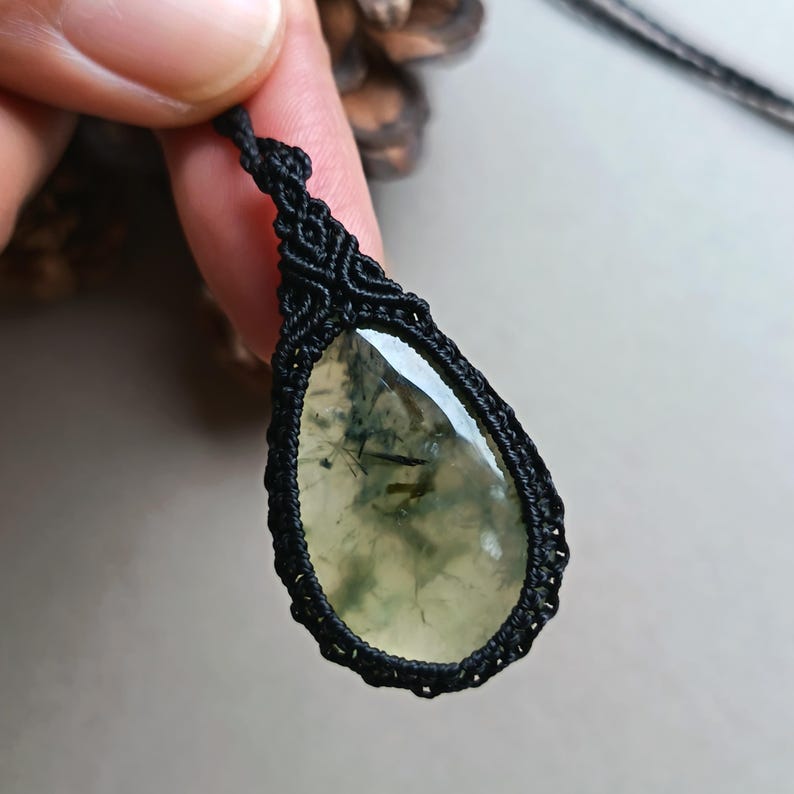 Op de afbeelding: Een druppelvormige hanger met een doorschijnende, lichtgroene steen, met donkergroene insluitsels. De steen is gevat in een zwarte macrame-omgeving en hangt aan een zwart koord. De hanger wordt door een hand omhoog gehouden.