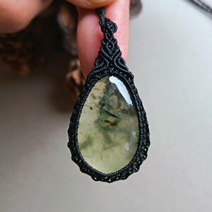 Op de afbeelding: Een druppelvormige hanger met een lichtgroene steen, met donkergroene insluitsels, is ingekapseld in een zwart macrame-ontwerp. De hanger hangt aan een zwart koord, waardoor een unieke en handgemaakte ketting ontstaat.