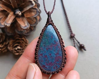 May include: A teardrop-shaped pendant necklace featuring a blue and red stone, encased in a brown macrame setting. The pendant hangs from a matching brown cord. The necklace is held in a hand, with a pinecone in the background.