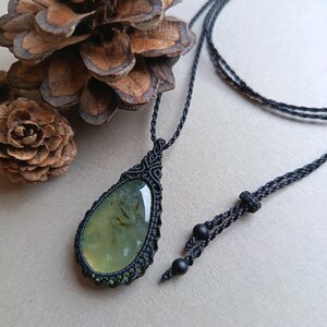 Op de afbeelding: Een handgemaakte macram&eacute; ketting met een druppelvormige, lichtgroene edelsteen hanger. De hanger is gevat in een ingewikkeld zwart macram&eacute; knoopontwerp, hangend aan een zwart koord. Twee zwarte kralen bevinden zich aan het einde van het verstelbare koord.