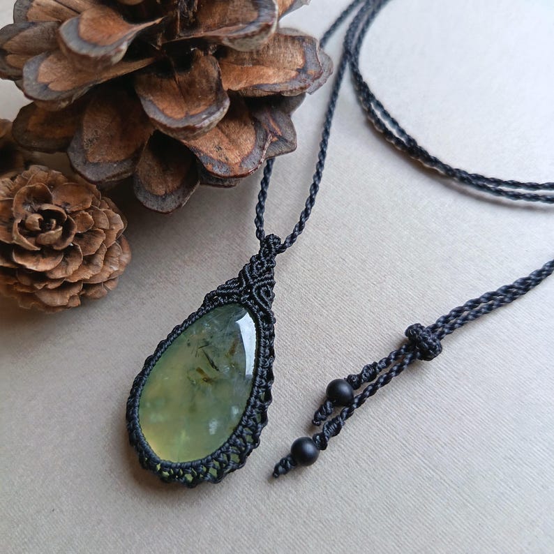 Op de afbeelding: Een handgemaakte macram&eacute; ketting met een druppelvormige, lichtgroene edelsteen hanger. De hanger is gevat in een ingewikkeld zwart macram&eacute; knoopontwerp, hangend aan een zwart koord. Twee zwarte kralen bevinden zich aan het einde van het verstelbare koord.