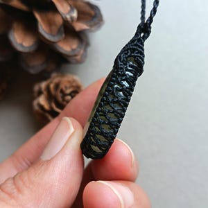 Op de afbeelding: Een close-up van een handgemaakte hangerketting. De hanger is een lichtgroene steen gewikkeld in een zwart macrame-ontwerp. Het kettingkoord is ook zwart. De achtergrond bevat een dennenappel.