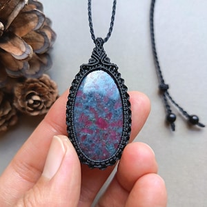 May include: A handmade macrame necklace featuring an oval-shaped pendant with a blue and red stone. The stone is set in a black macrame frame and hangs from a black cord. The pendant is approximately 5 cm long.