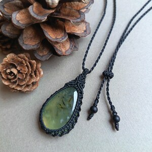 Op de afbeelding: Een handgemaakte macram&eacute; ketting met een druppelvormige, lichtgroene edelsteen hanger. De hanger is omhuld in ingewikkeld zwart macram&eacute;, hangend aan een zwart koord met verstelbare zwarte kralen. De ketting ligt op een neutrale ondergrond met dennenappels.