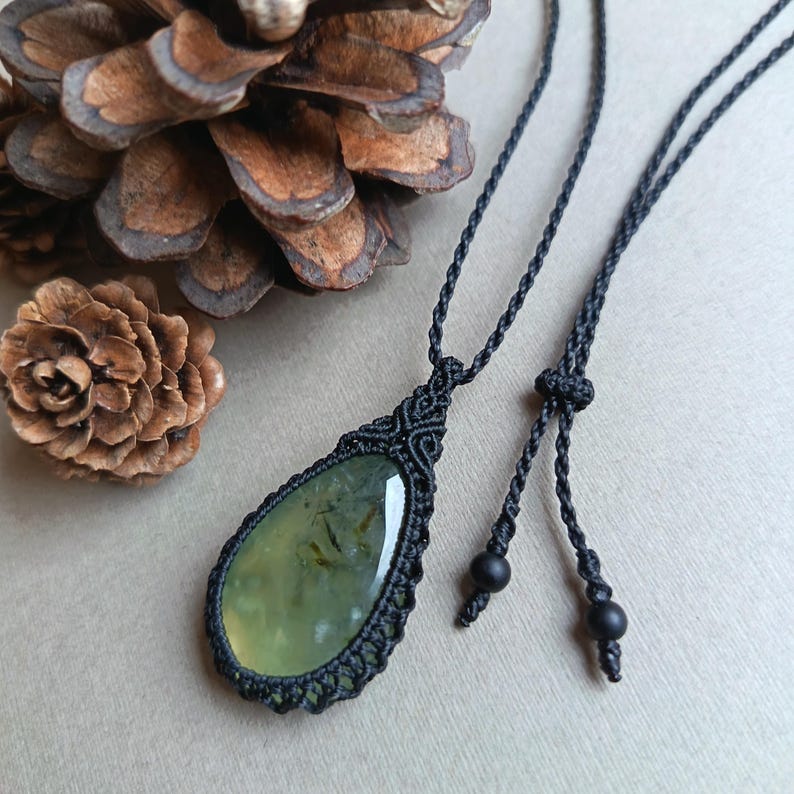 Op de afbeelding: Een handgemaakte macram&eacute; ketting met een druppelvormige, lichtgroene edelsteen hanger. De hanger is omhuld in ingewikkeld zwart macram&eacute;, hangend aan een zwart koord met verstelbare zwarte kralen. De ketting ligt op een neutrale ondergrond met dennenappels.