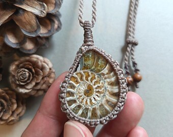 May include: A pendant featuring a spiral-shaped ammonite fossil with amber and brown tones, encased in a light brown macrame cord setting. The necklace includes a matching adjustable cord with two small wooden beads at the ends for a secure fit.