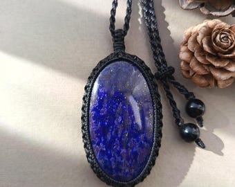 May include: An oval-shaped lapis lazuli pendant featuring deep blue tones with gold-colored flecks, encased in a black macrame cord setting. The pendant hangs from a matching black braided cord that includes two small black beads at the end.