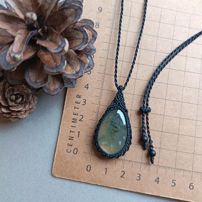 Op de afbeelding: Een zwarte macram&eacute; ketting met een druppelvormige hanger met een lichtgroene steen. De hanger wordt getoond op een bruin oppervlak met een centimeterliniaal. Een dennenappel is op de achtergrond.