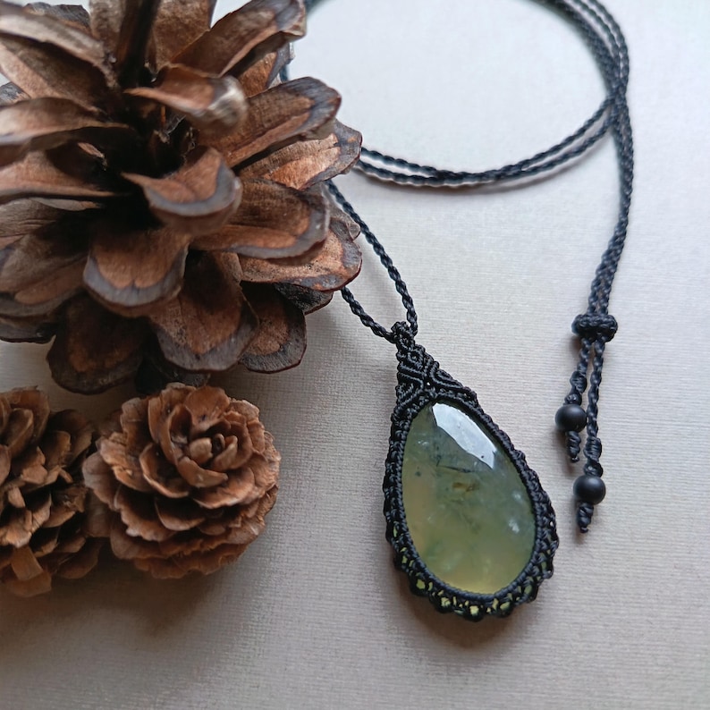 Op de afbeelding: Een handgemaakte macram&eacute; ketting met een peervormige, lichtgroene edelsteen hanger. De hanger is omhuld met ingewikkeld zwart macram&eacute;, hangend aan een bijpassend zwart koord. De achtergrond bevat dennenappels.