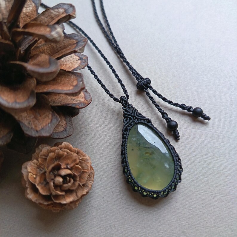 Op de afbeelding: Een handgemaakte macram&eacute; ketting met een druppelvormige, lichtgroene edelsteen hanger. De hanger is gevat in ingewikkeld zwart macram&eacute;, hangend aan een bijpassend zwart koord. De ketting wordt getoond op een neutrale ondergrond met dennenappels.