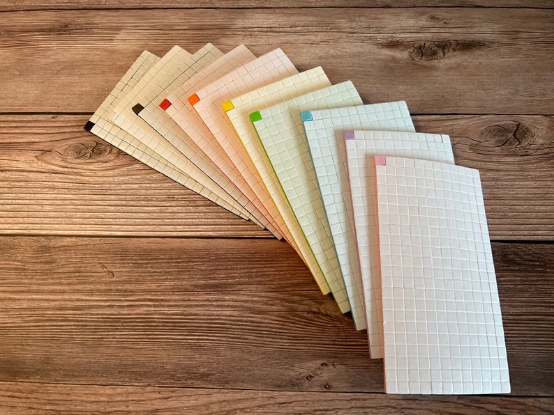 May include: A set of ten gridded notebooks with colorful accents. The notebooks are arranged in a fan shape, with the covers facing outward. The notebooks are all the same size and have a white background with a grid pattern.