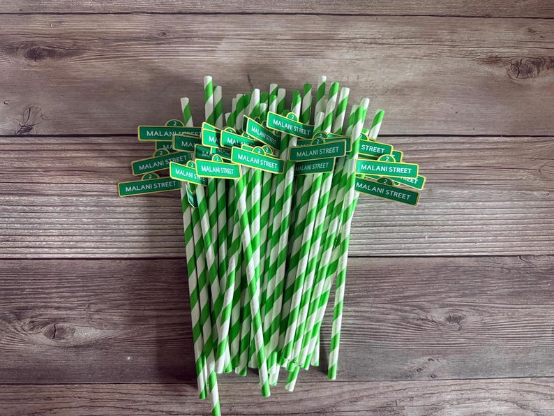 May include: A bundle of green and white striped paper straws, each adorned with a small sign that reads "MALANI STREET" in white text on a green background. The straws are arranged against a wooden background.