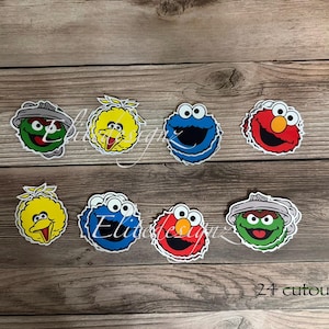 May include: A collection of colorful character cutouts, including Big Bird, Cookie Monster, Elmo, and Oscar the Grouch, arranged on a wooden surface. The image includes the text "24 cutouts".