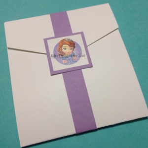 May include: A white envelope with a purple stripe down the center. A square sticker with a cartoon image of a princess with brown hair and a blue dress is on the envelope. The text "Elite Designs by Zen" is on the sticker.