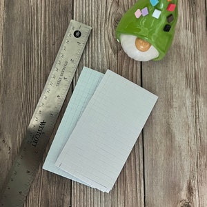 May include: Two sheets of white grid paper with a ruler on a wooden surface. The ruler is made of stainless steel and has markings in inches. A green gnome with a white face and colorful squares on its hat is in the upper right corner of the image.