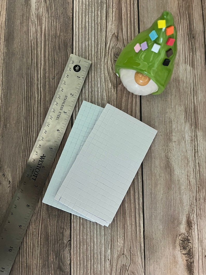 May include: Two sheets of white grid paper with a ruler on a wooden surface. The ruler is made of stainless steel and has markings in inches. A green gnome with a white face and colorful squares on its hat is in the upper right corner of the image.