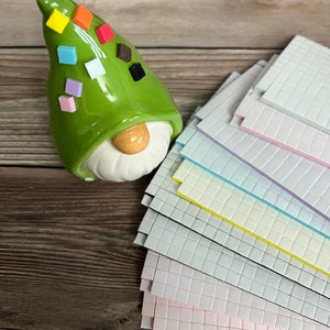 May include: A green ceramic gnome figurine with a hat decorated with colorful square tiles. The gnome is sitting on a wooden surface next to a stack of colorful square tiles.