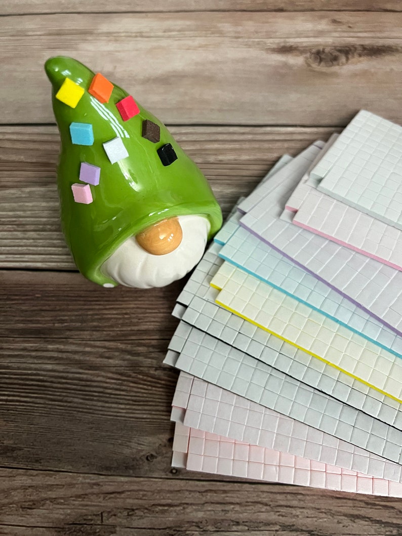 May include: A green ceramic gnome figurine with a hat decorated with colorful square tiles. The gnome is sitting on a wooden surface next to a stack of colorful square tiles.