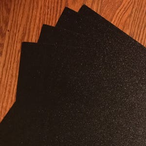 May include: A stack of black glitter paper sheets. The paper has a fine, sparkly texture.