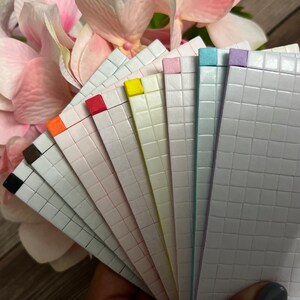 May include: A set of ten adhesive-backed, gridded paper sheets in various pastel colors, including black, brown, orange, red, yellow, green, blue, and purple. The sheets are arranged in a fan shape, with the colored edges facing outward.