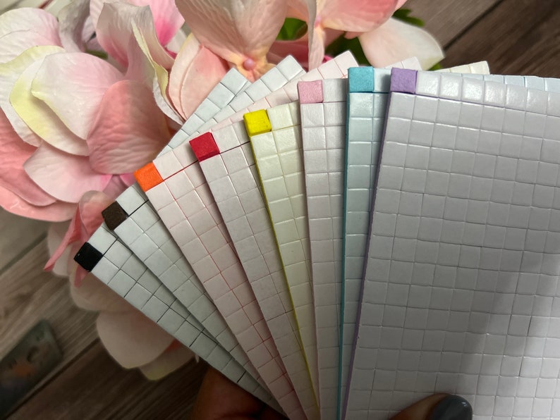 May include: A set of ten adhesive-backed, gridded paper sheets in various pastel colors, including black, brown, orange, red, yellow, green, blue, and purple. The sheets are arranged in a fan shape, with the colored edges facing outward.