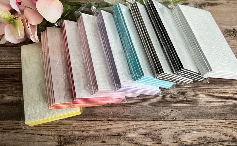May include: A variety pack of adhesive foam squares in different colors, including yellow, orange, red, pink, purple, blue, green, black, and white. The squares are packaged in clear plastic and are stacked on a wooden surface.