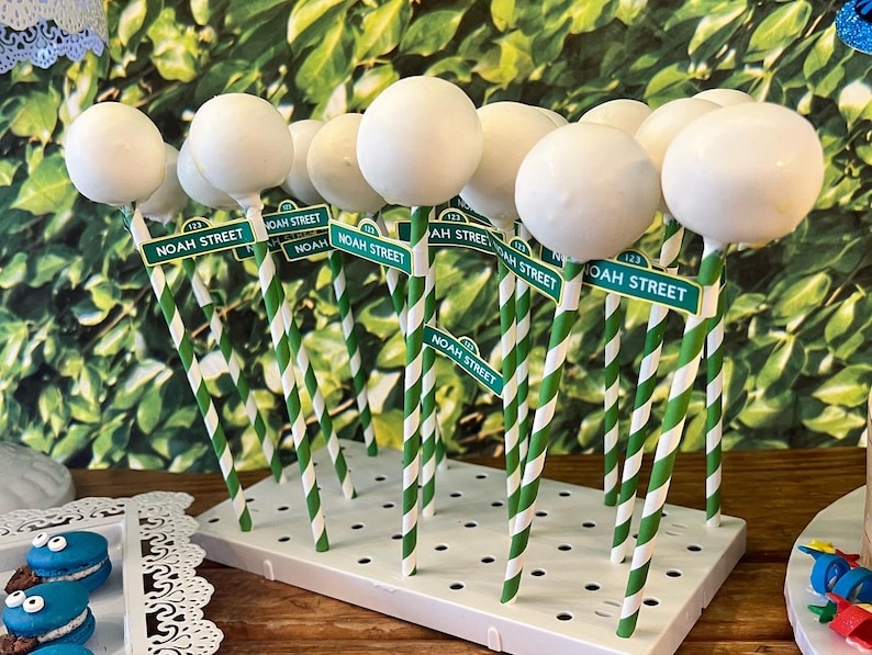May include: White cake pops on green and white striped sticks, each with a small sign that reads "NOAH STREET". The cake pops are arranged on a white display stand, with a green leafy background.