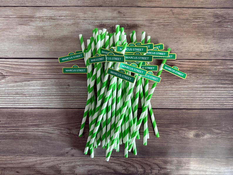 May include: A bundle of green and white striped paper straws with small green street sign toppers that read "MARCUS STREET". The straws are arranged on a wooden surface.