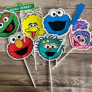 May include: A collection of Sesame Street character cutouts on sticks, including Elmo, Cookie Monster, Big Bird, and others. A green sign that reads "Noah Street" is also included. A blue glittery number one is also included.