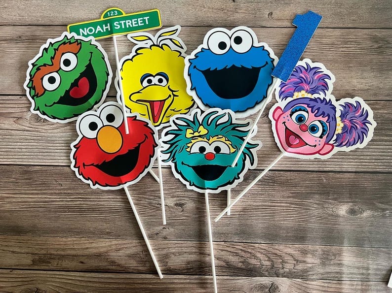 May include: A collection of Sesame Street character cutouts on sticks, including Elmo, Cookie Monster, Big Bird, and others. A green sign that reads "Noah Street" is also included. A blue glittery number one is also included.
