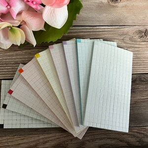 May include: A set of ten gridded paper sheets in various colors, including red, orange, yellow, green, blue, purple, and white. The sheets are arranged in a fan shape on a wooden surface.