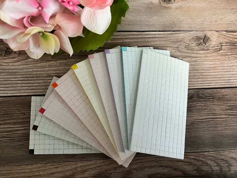 May include: A set of ten gridded paper sheets in various colors, including red, orange, yellow, green, blue, purple, and white. The sheets are arranged in a fan shape on a wooden surface.