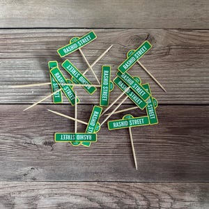 May include: Green and white "Rashid Street" sign cupcake toppers with wooden toothpicks. The toppers are scattered on a wooden surface, ideal for themed parties or events.