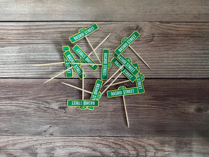 May include: Green and white "Rashid Street" sign cupcake toppers with wooden toothpicks. The toppers are scattered on a wooden surface, ideal for themed parties or events.