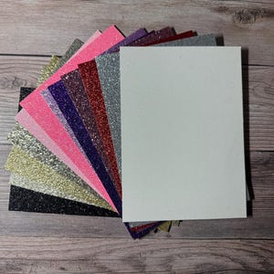 May include: A collection of glitter cardstock sheets in various colors, including black, gold, pink, purple, and silver. A large white cardstock sheet is also included. The sheets are arranged in a fan shape, showcasing the different colors and textures.