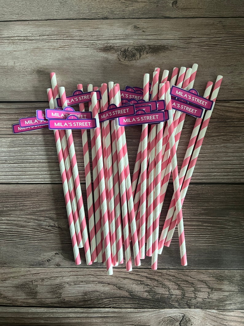 May include: A bundle of pink and white striped paper straws. Each straw has a small sign that reads "Mila's Street" in white text on a purple background. The straws are arranged on a wooden surface.