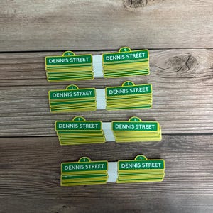 May include: Stacks of green and yellow "DENNIS STREET" signs with a white backing. Each sign has a green top with the number "1" and a green and yellow rectangular sign with the street name. The signs are on a wooden surface.