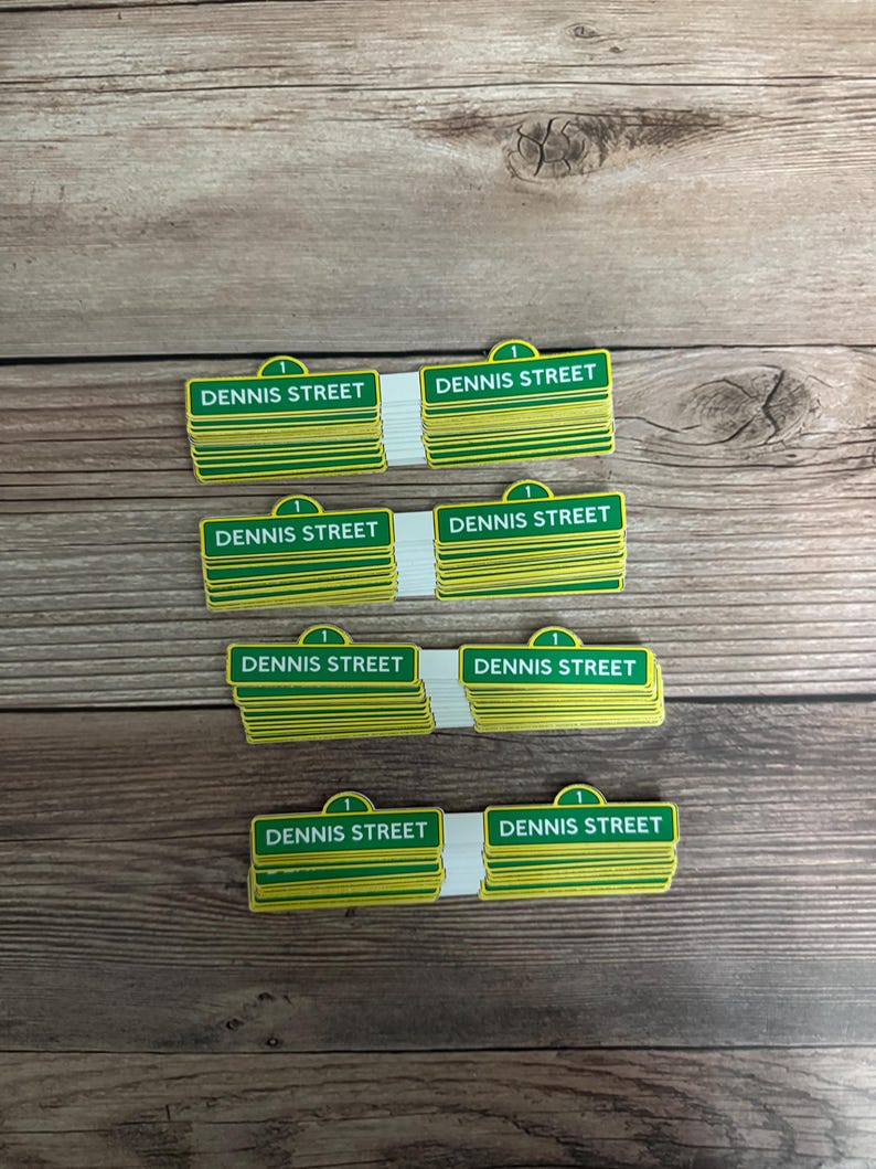 May include: Stacks of green and yellow "DENNIS STREET" signs with a white backing. Each sign has a green top with the number "1" and a green and yellow rectangular sign with the street name. The signs are on a wooden surface.